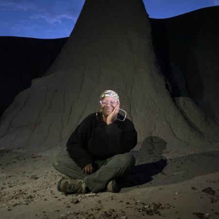 Siesa is sitting cross legged on the ground in front of a smooth rock formation at dusk, and wearing a light toned head scarf, hoop earrings, a black sweater and dark green pants